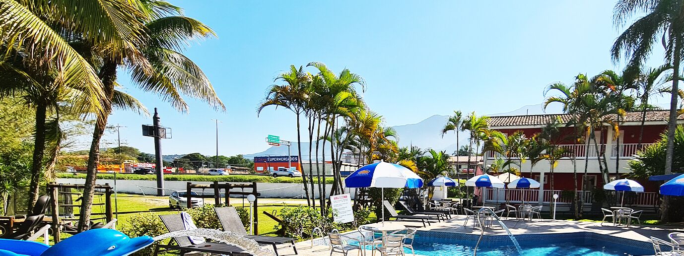 Zona de piscina con sillones de descanso y sombrillas, palmeras y vista a una carretera con un supermercado al fondo.