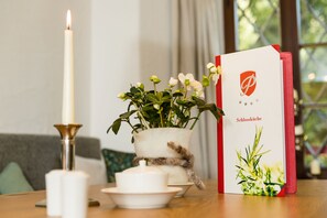 Una mesa con una vela encendida, una planta en maceta, una tetera blanca y una tarjeta roja y blanca con un logotipo y texto.
