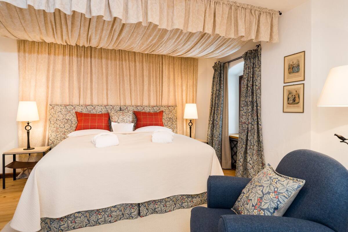 A hotel room with a large bed, two bedside lamps, a blue armchair, and floral curtains.