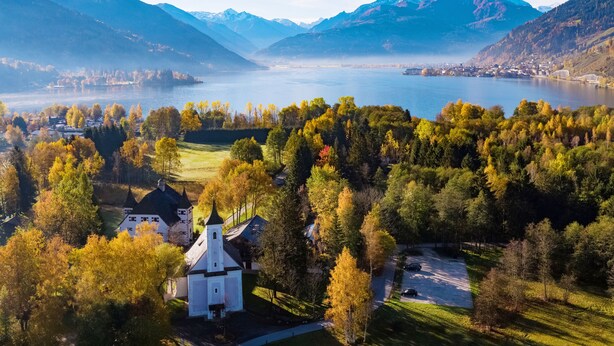 A scenic view of a lake surrounded by mountains and autumn-colored trees, with a small white church in the foreground. - Schloss Prielau Hotel & Restaurants (Zell am See)