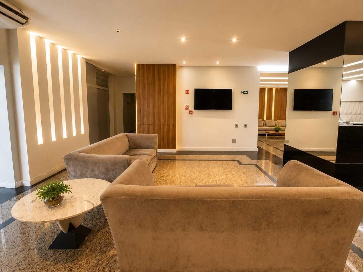 A modern hotel lobby with a seating area, a flat-screen TV mounted on the wall, and a potted plant on a round table.
