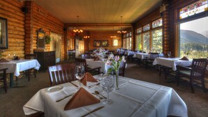 A log cabin-style dining area with multiple tables set for dining, large windows with mountain views, and wooden walls. - Overlander Mountain Lodge (Jasper East)
