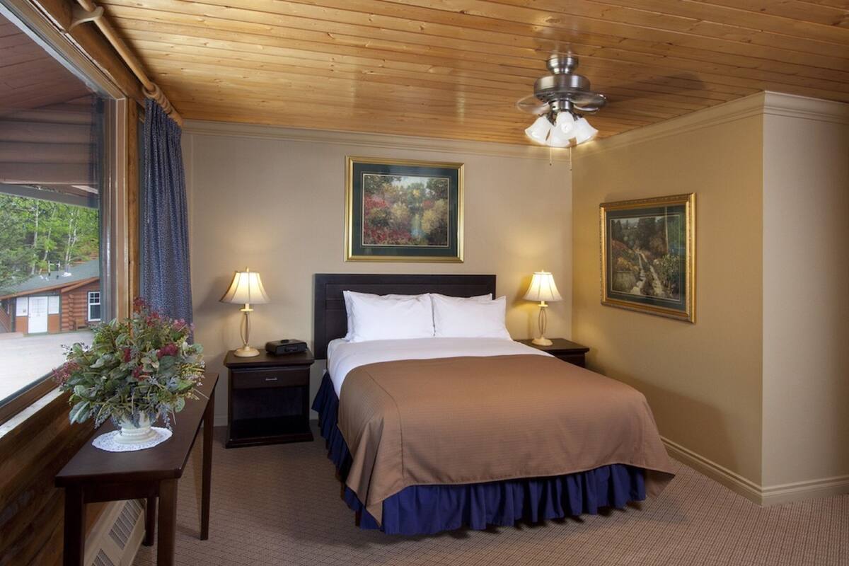 A hotel room with a bed, two bedside tables with lamps, a window with curtains, and two framed pictures on the wall.
