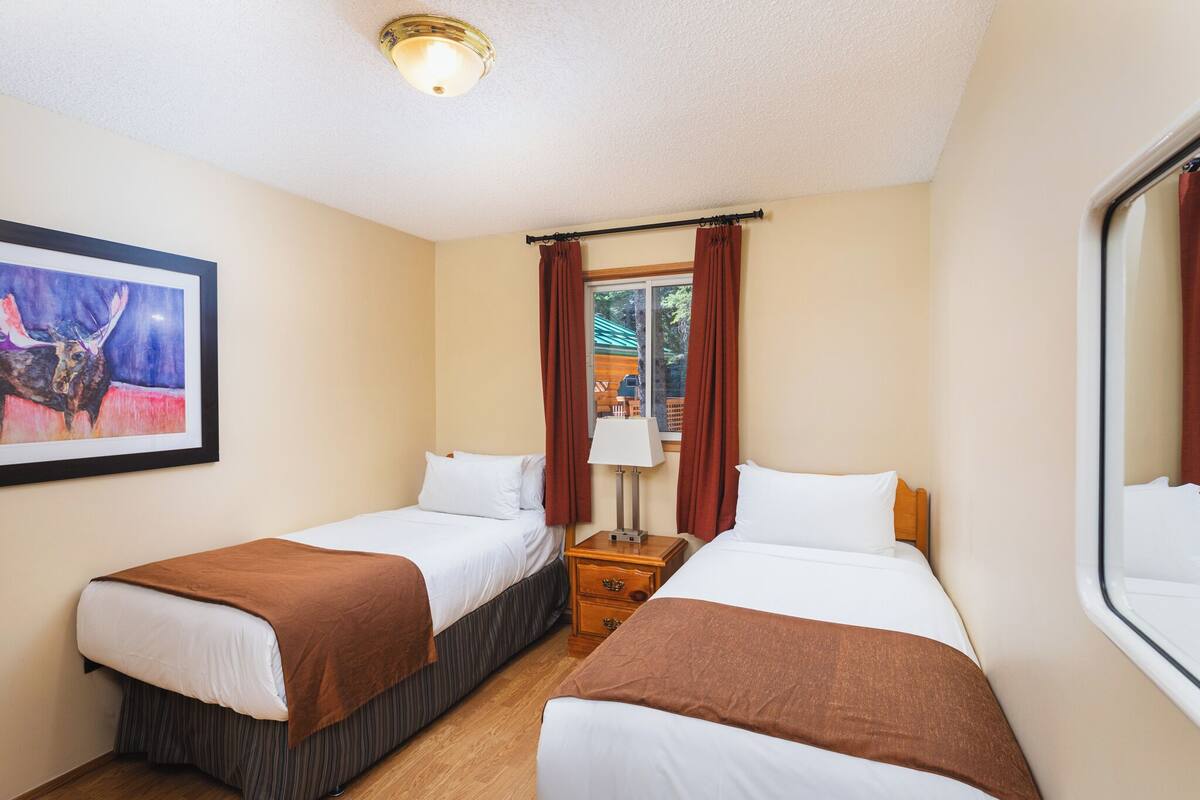 A hotel room with two single beds, a nightstand with a lamp, a framed picture on the wall, and a window with curtains.