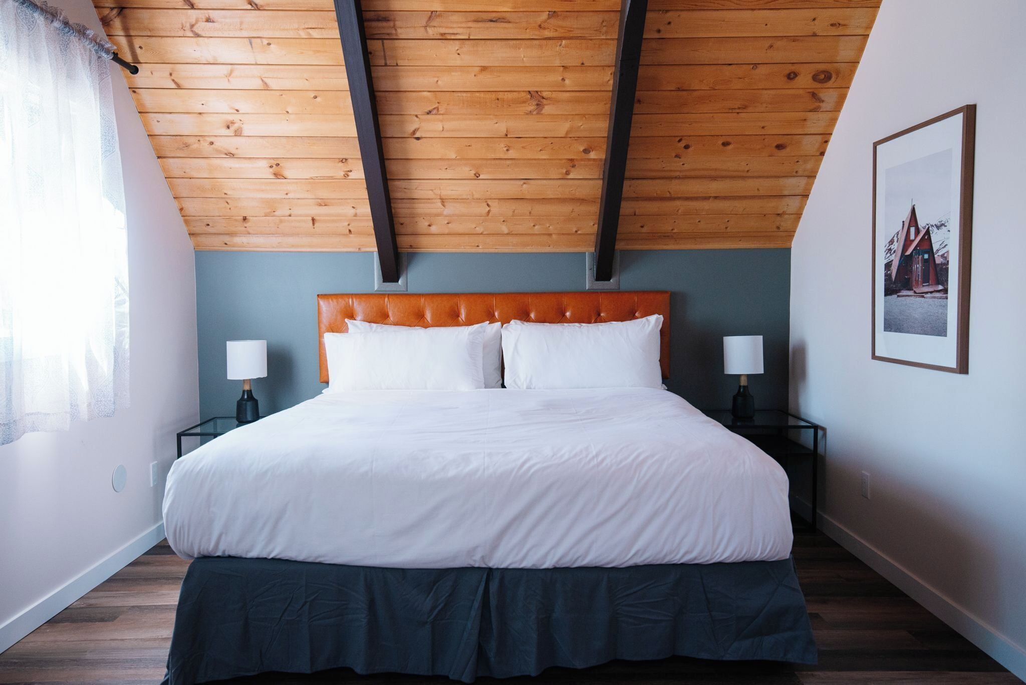 Une chambre avec un plafond en bois, un grand lit avec une literie blanche, deux tables de chevet avec des lampes et un tableau encadré au mur.