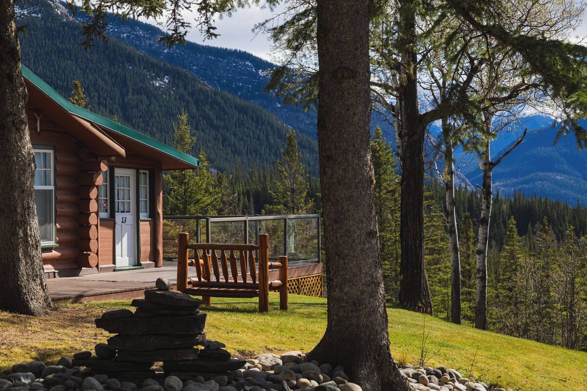A log cabin with a green roof, a wooden bench, and a stone stack in a mountainous area.