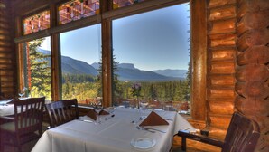 A dining area with a table set for two, large windows offering a mountain view, and wooden log walls. - Overlander Mountain Lodge (Jasper East)