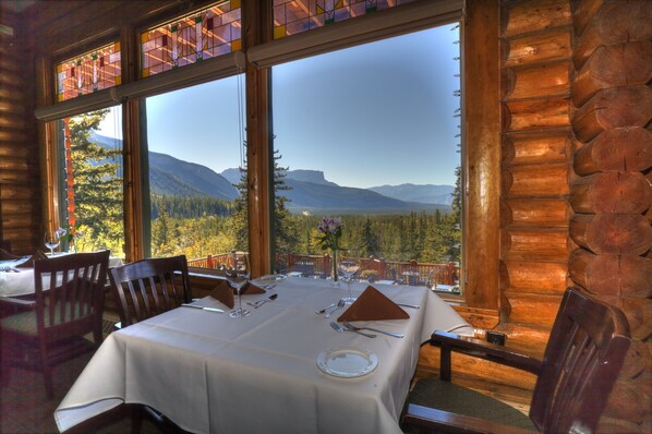 A dining area with a table set for two, large windows offering a mountain view, and wooden log walls.