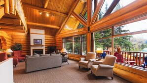 A log cabin interior with a fireplace, seating area, and large windows offering a view of the outdoors. - Overlander Mountain Lodge (Jasper East)