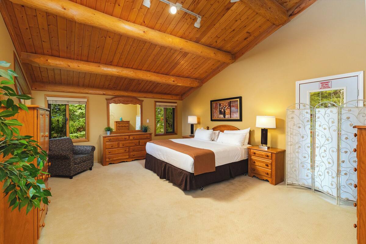 A bedroom with a wooden ceiling, a large bed, a dresser, a chair, and a window.