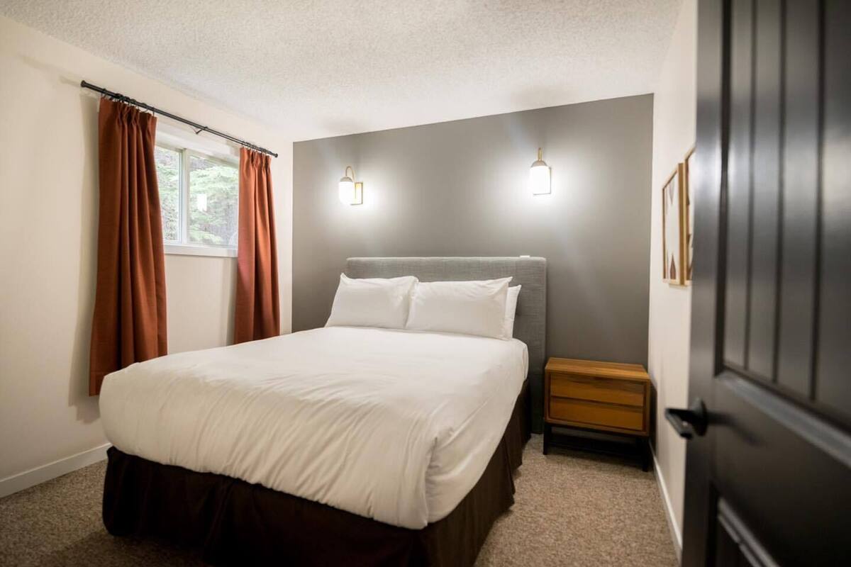 A bedroom with a bed, a nightstand, a window with curtains, and wall-mounted lighting.