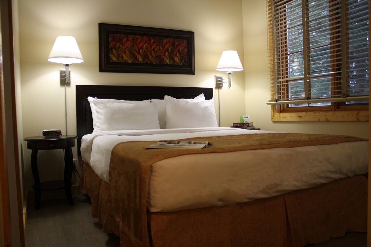 A hotel room with a bed, two bedside lamps, a nightstand, a painting on the wall, and a window with blinds.