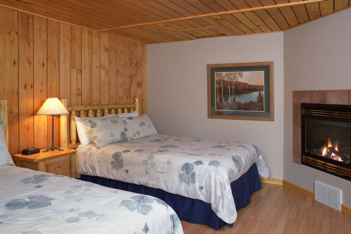 A cozy room with two beds, a wooden ceiling, a fireplace, and a framed picture on the wall.