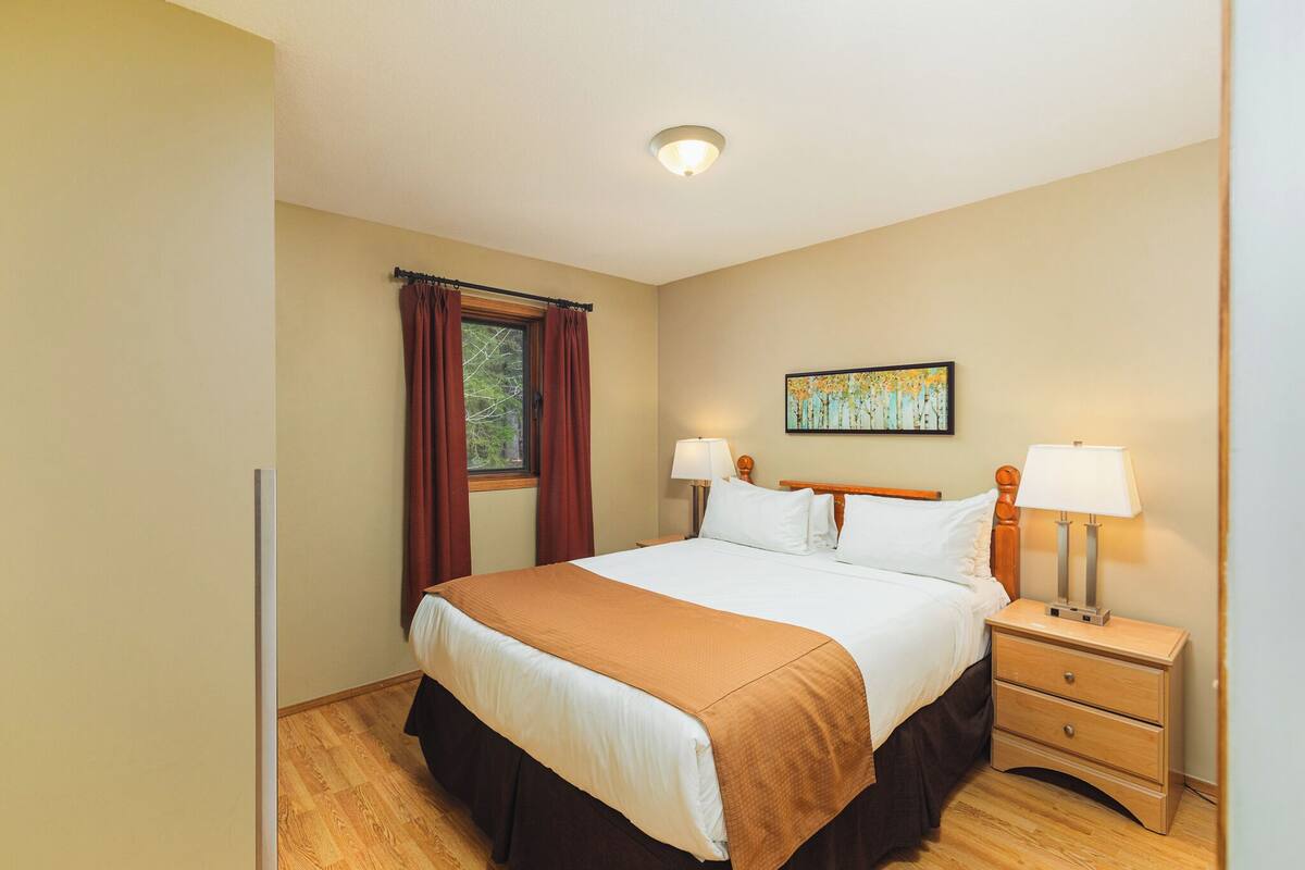 A neatly arranged bedroom with a large bed, two bedside tables with lamps, a window with curtains, and a painting on the wall.