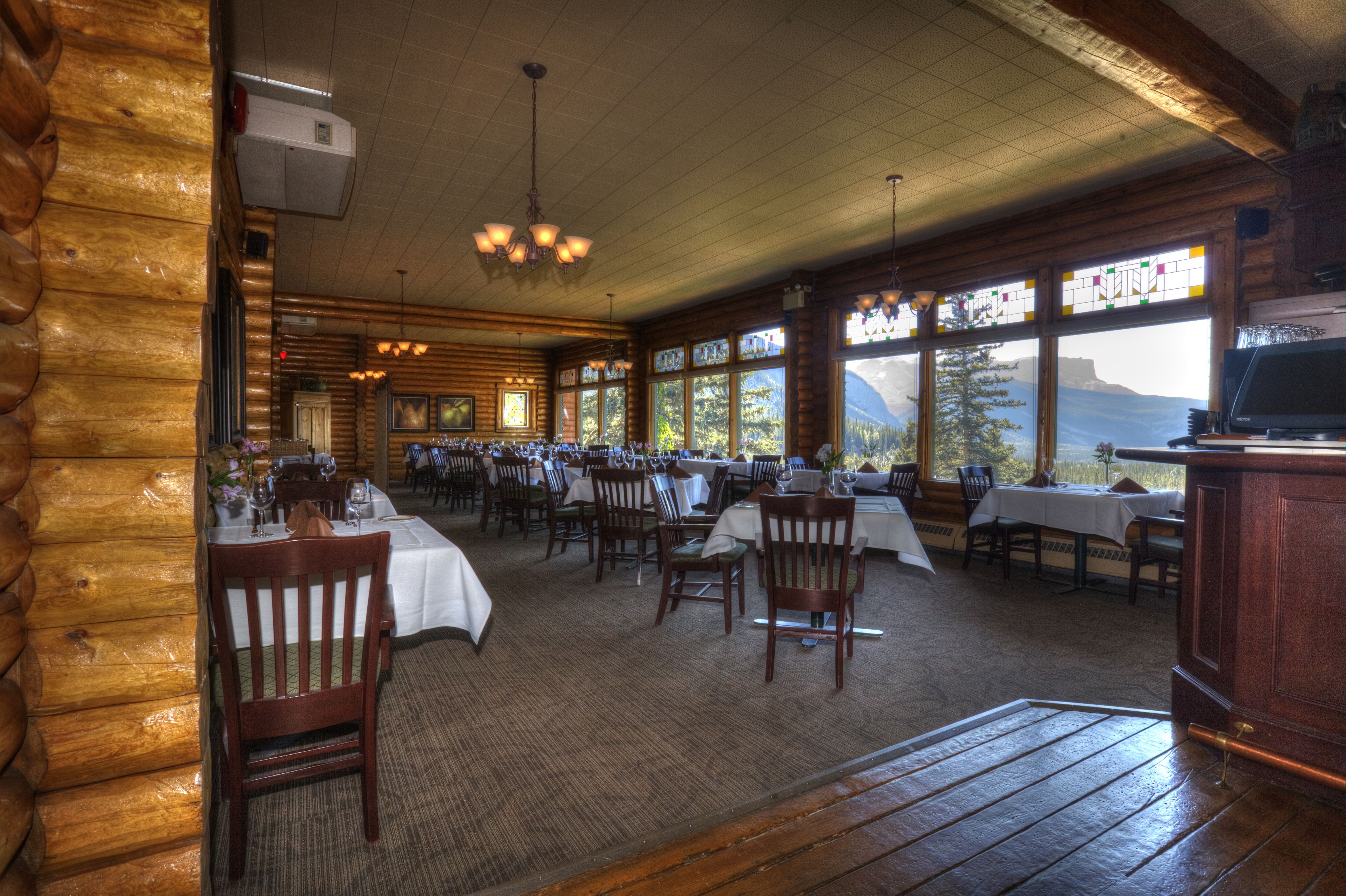 A log cabin-style restaurant with multiple dining tables and chairs, large windows with mountain views, and wooden interior.