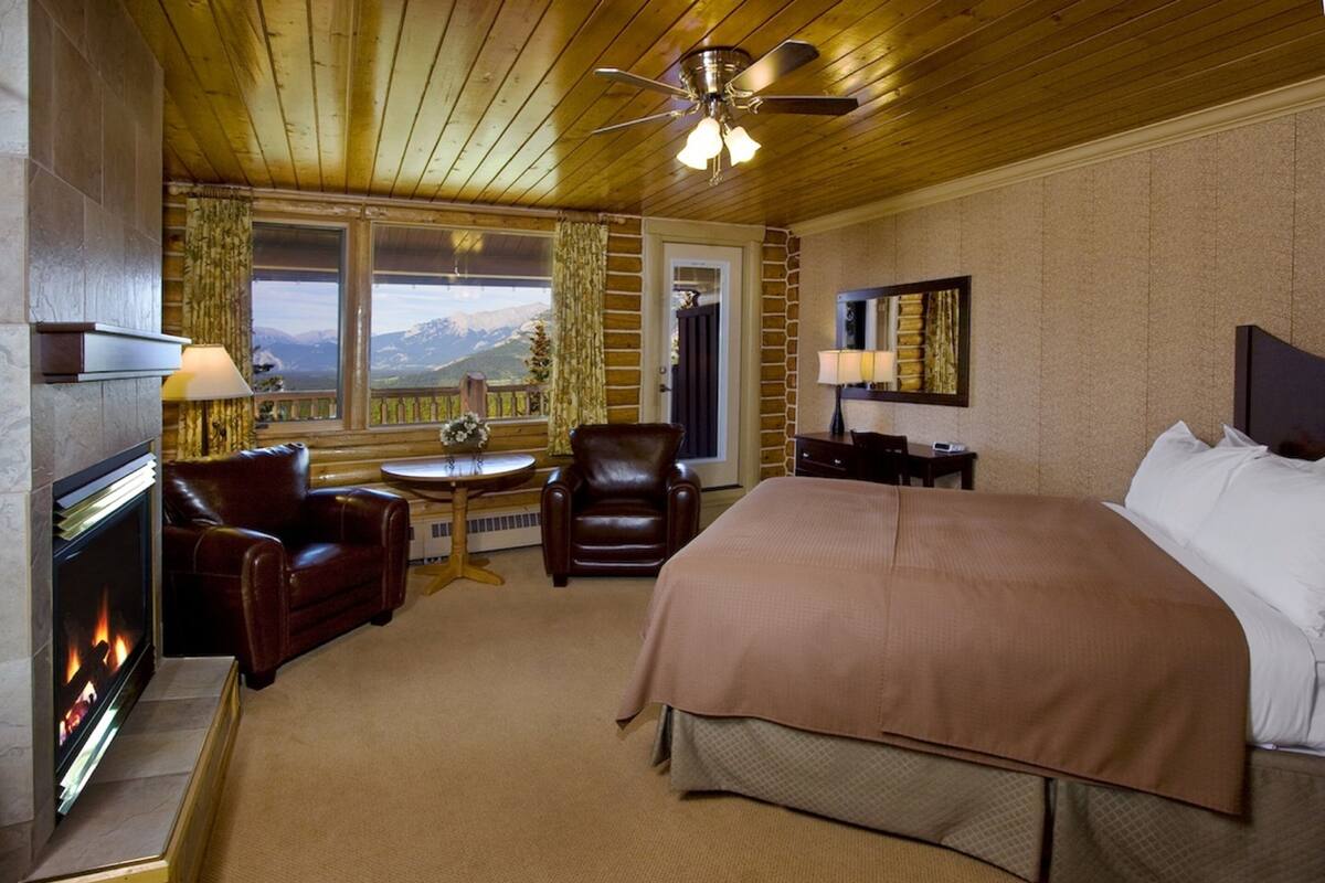 A hotel room with a large bed, a fireplace, leather chairs, a small table, and a view of mountains.