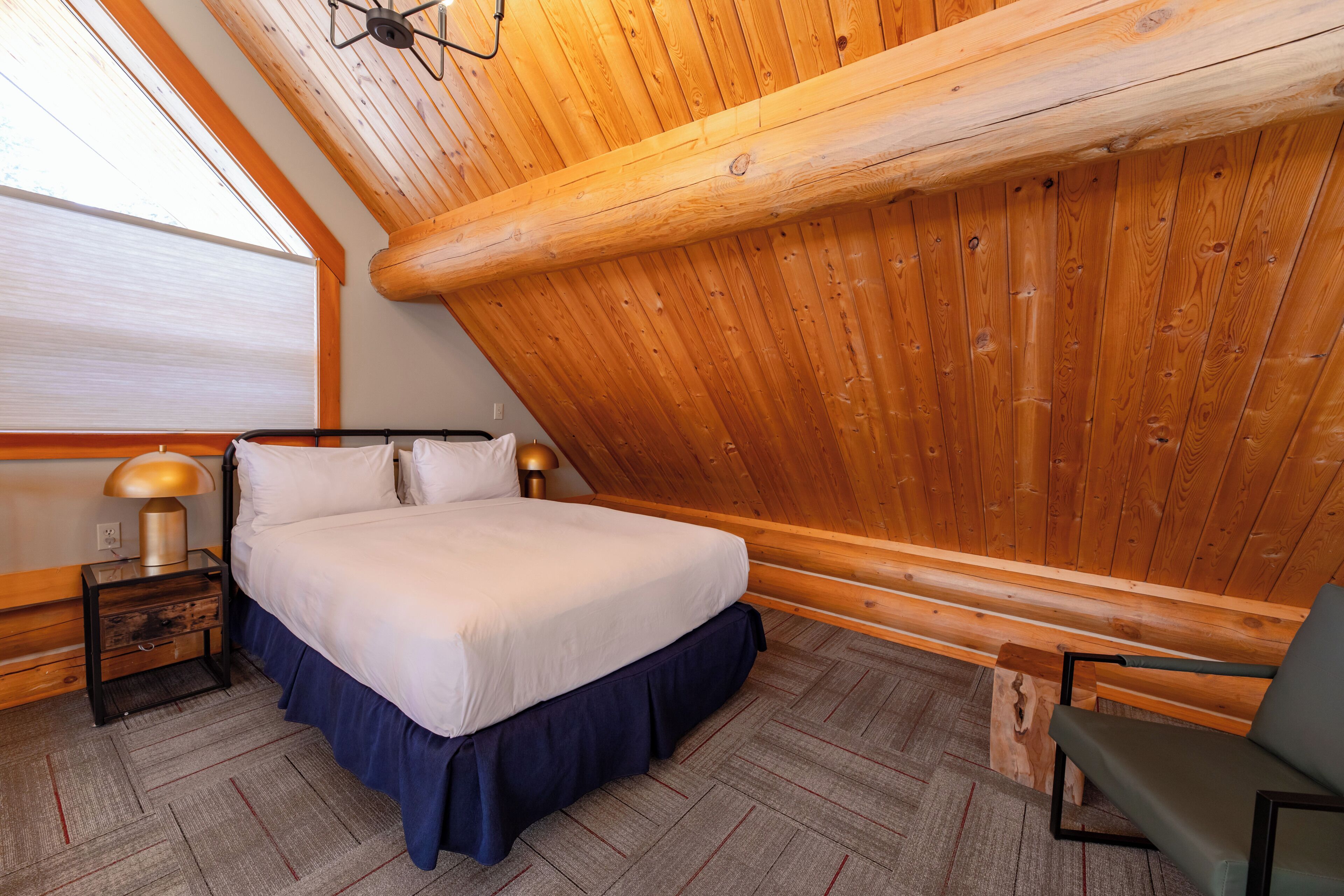 A bedroom with a wooden ceiling, a bed with white bedding, a nightstand with a lamp, and a chair.
