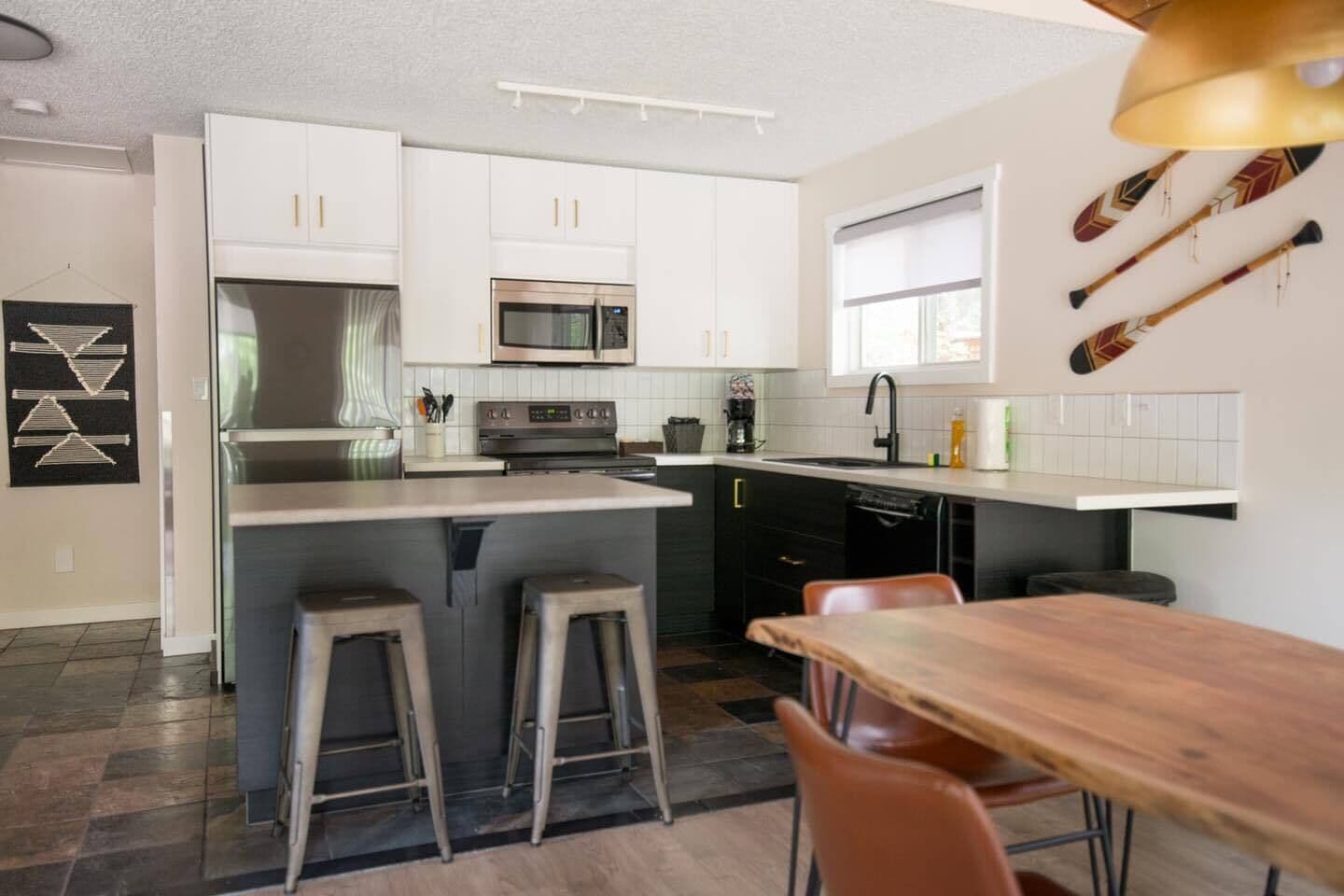Une cuisine moderne avec des meubles blancs, un îlot central doté de tabourets, une table à manger et une fenêtre avec des stores.