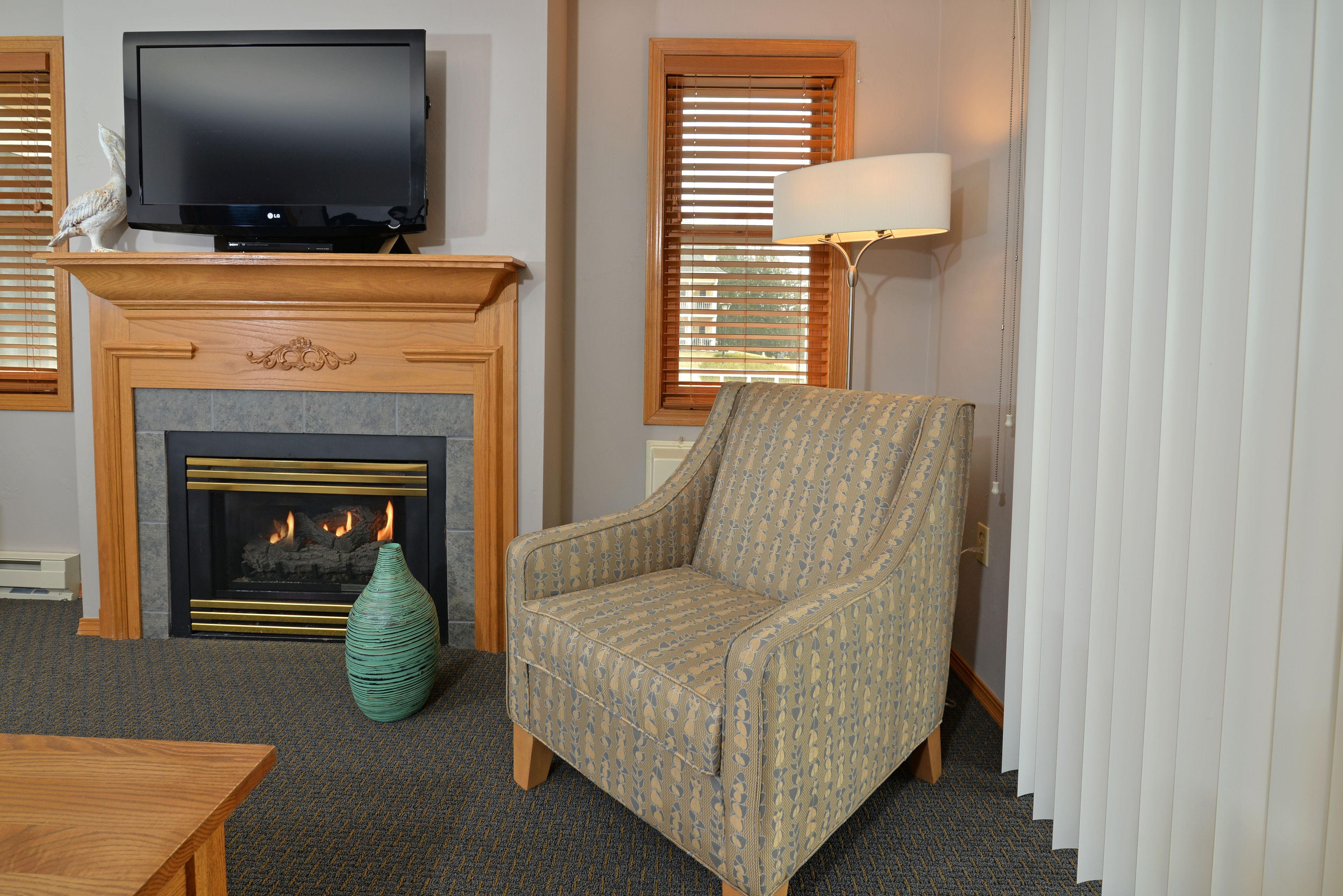 Una sala de estar con chimenea, televisor, un sillón estampado, un jarrón verde y una lámpara.