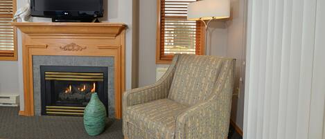 Una sala de estar con chimenea, televisor, un sillón estampado, un jarrón verde y una lámpara.