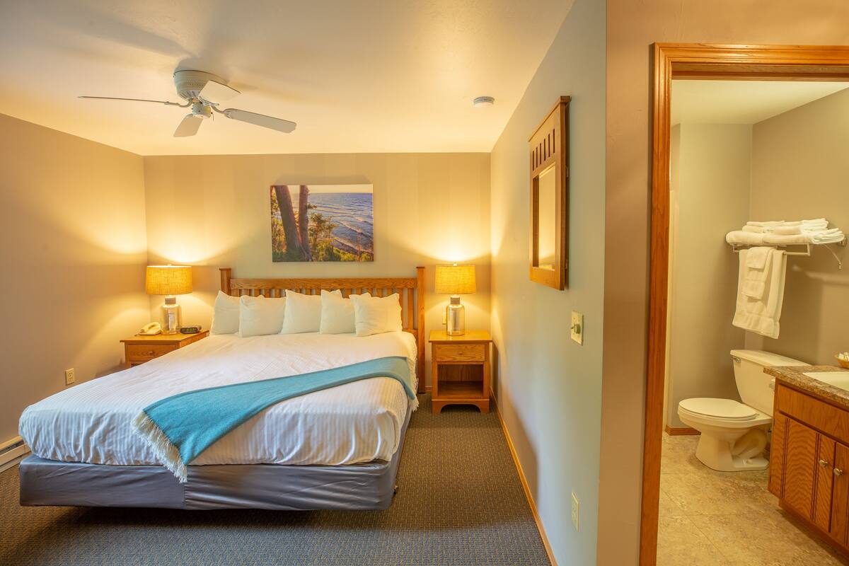 Una habitación de hotel con una cama, un ventilador de techo, dos lámparas de noche, un cuadro en la pared y un baño con inodoro y lavamanos.