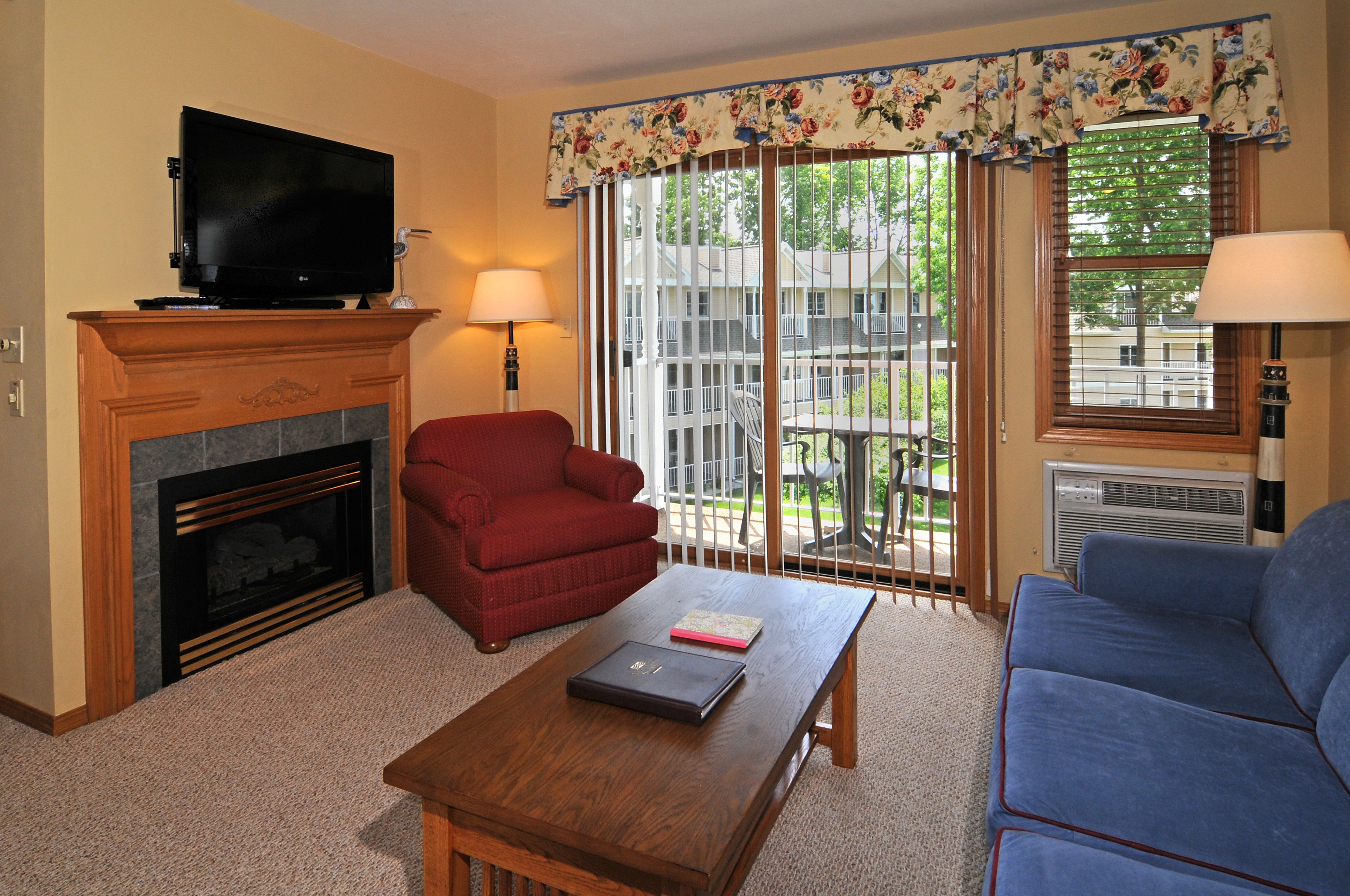 A living room with a fireplace, a television, a red armchair, a blue sofa, and a balcony with a view of a house and trees.