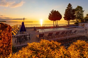 A poolside area with chairs and tables, a lamp post, and a view of the sunset over the water. - Westwood Shores Waterfront Resort (Sturgeon Bay)