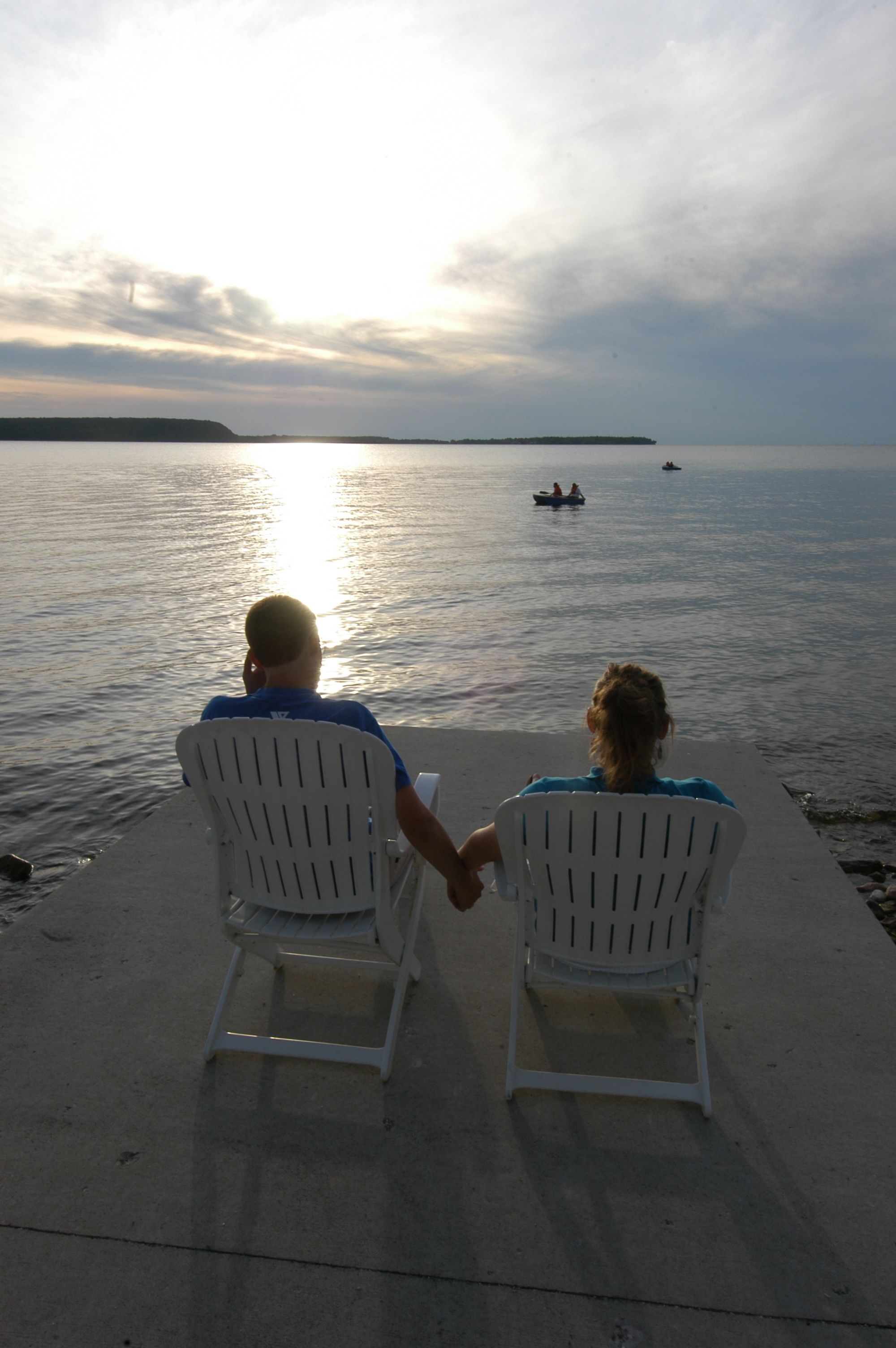 Two people sitting on white chairs facing a body of water with boats in the distance.