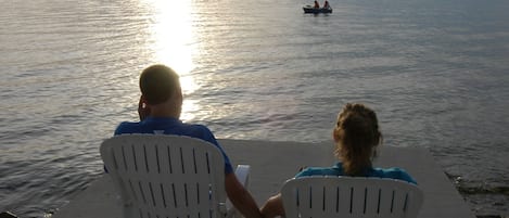 Two people sitting on white chairs facing a body of water with boats in the distance.
