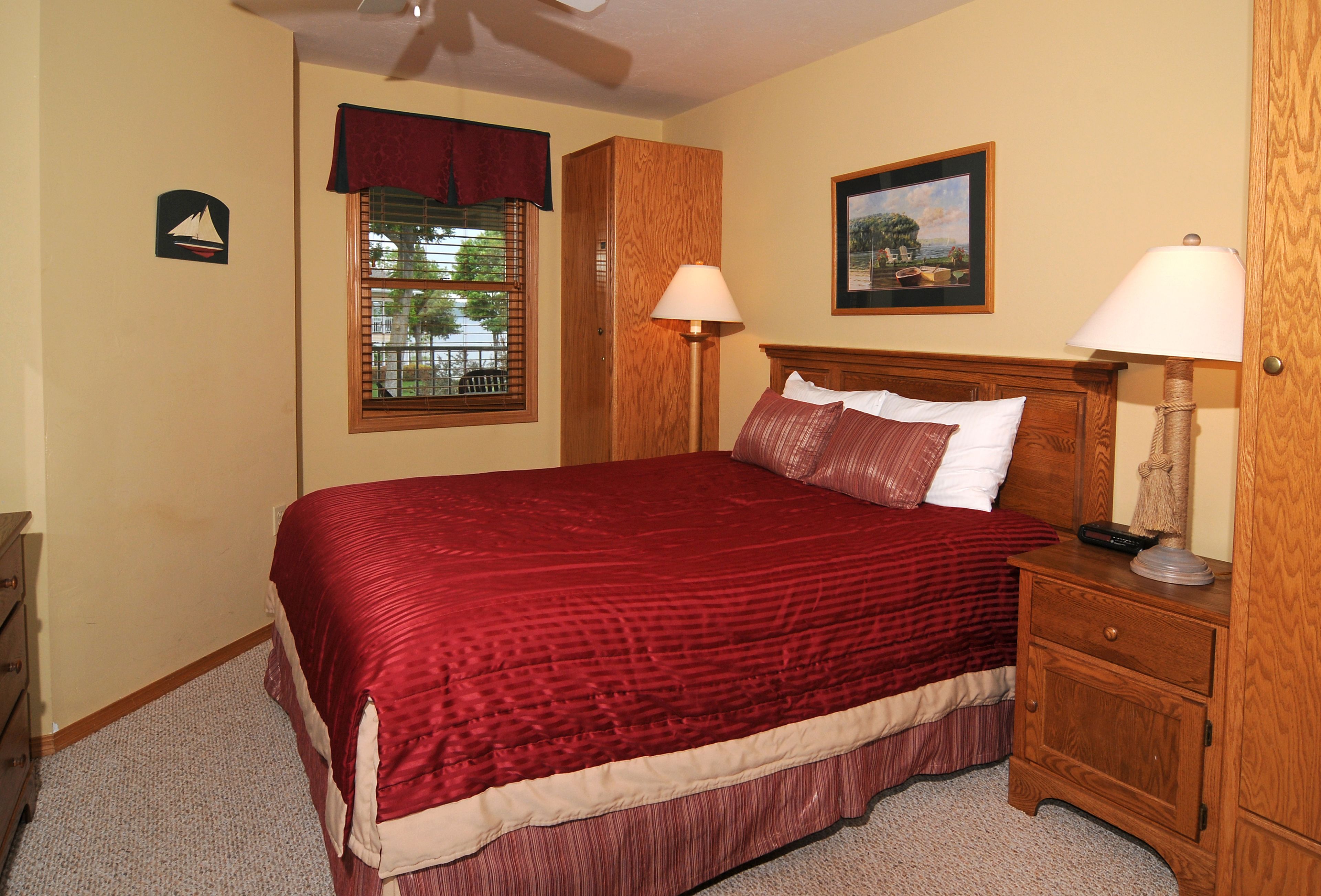 A bedroom with a large bed, two bedside tables, a nightstand, a window with blinds, a ceiling fan, and a framed picture on the wall.