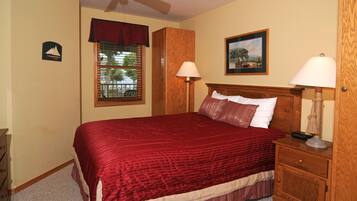A bedroom with a large bed, two bedside tables, a nightstand, a window with blinds, a ceiling fan, and a framed picture on the wall.