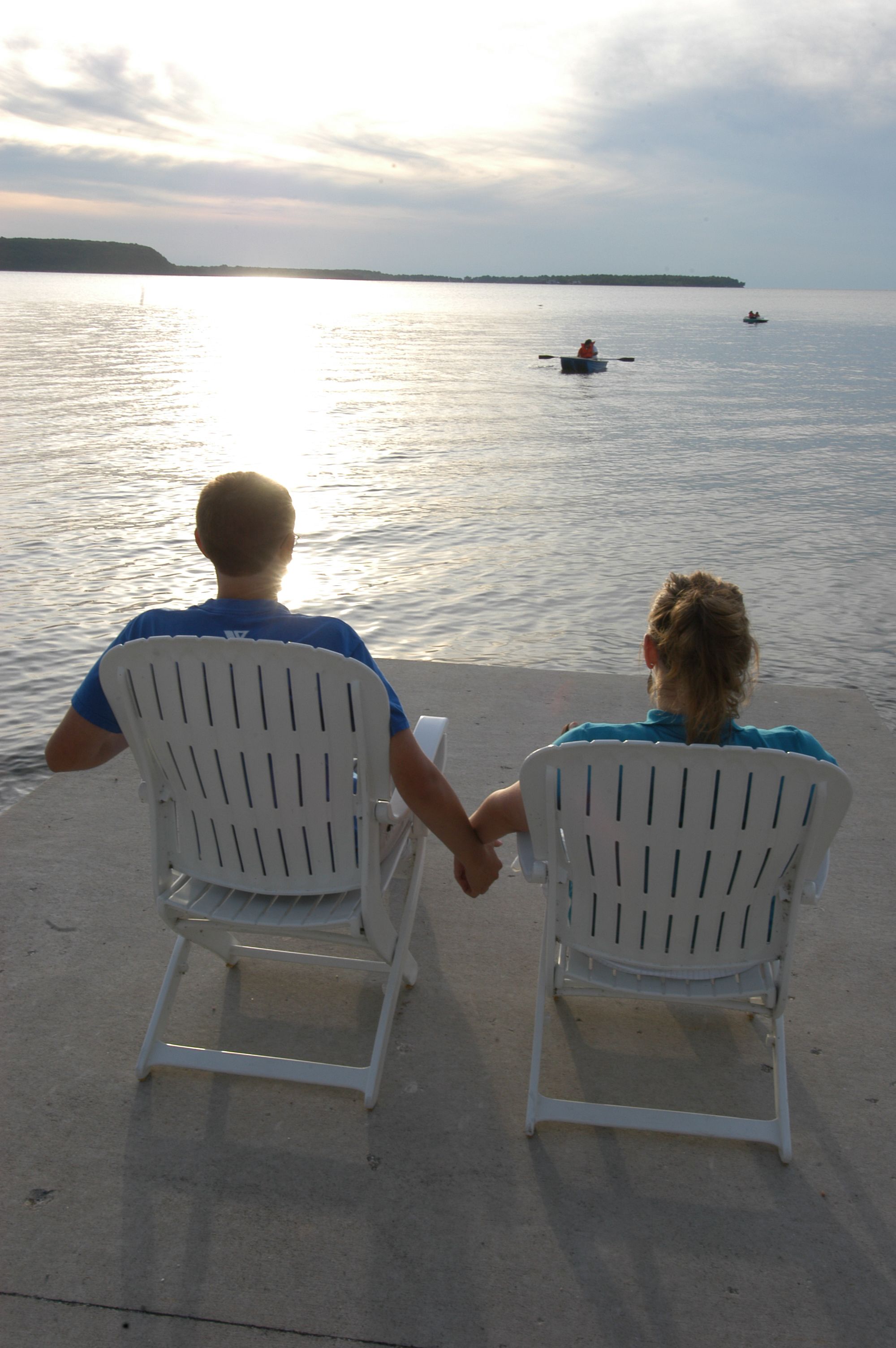 Two people seated on white chairs facing a body of water with a distant shoreline.