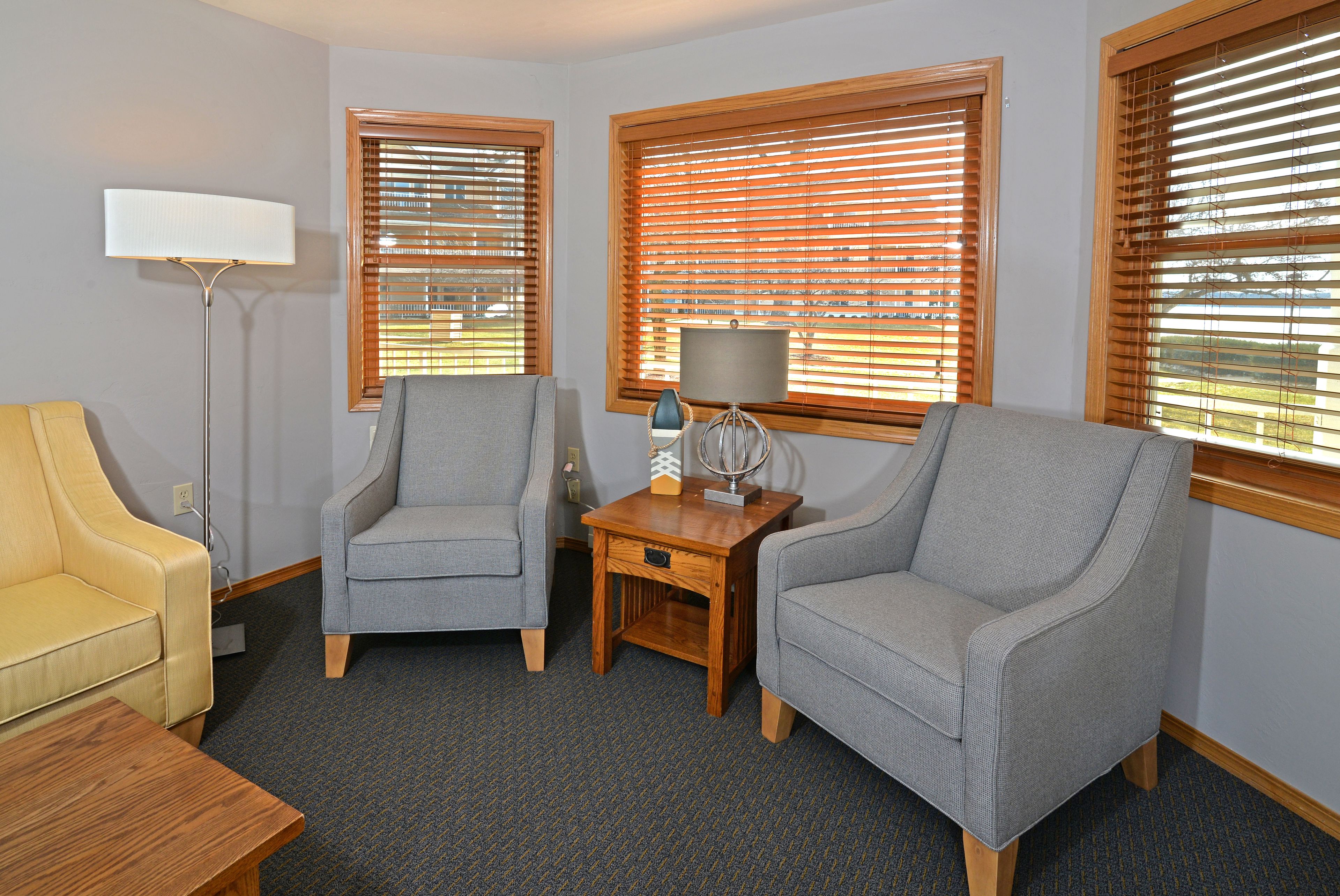 A living room with two grey armchairs, a yellow sofa, a wooden side table with a lamp, and a floor lamp.