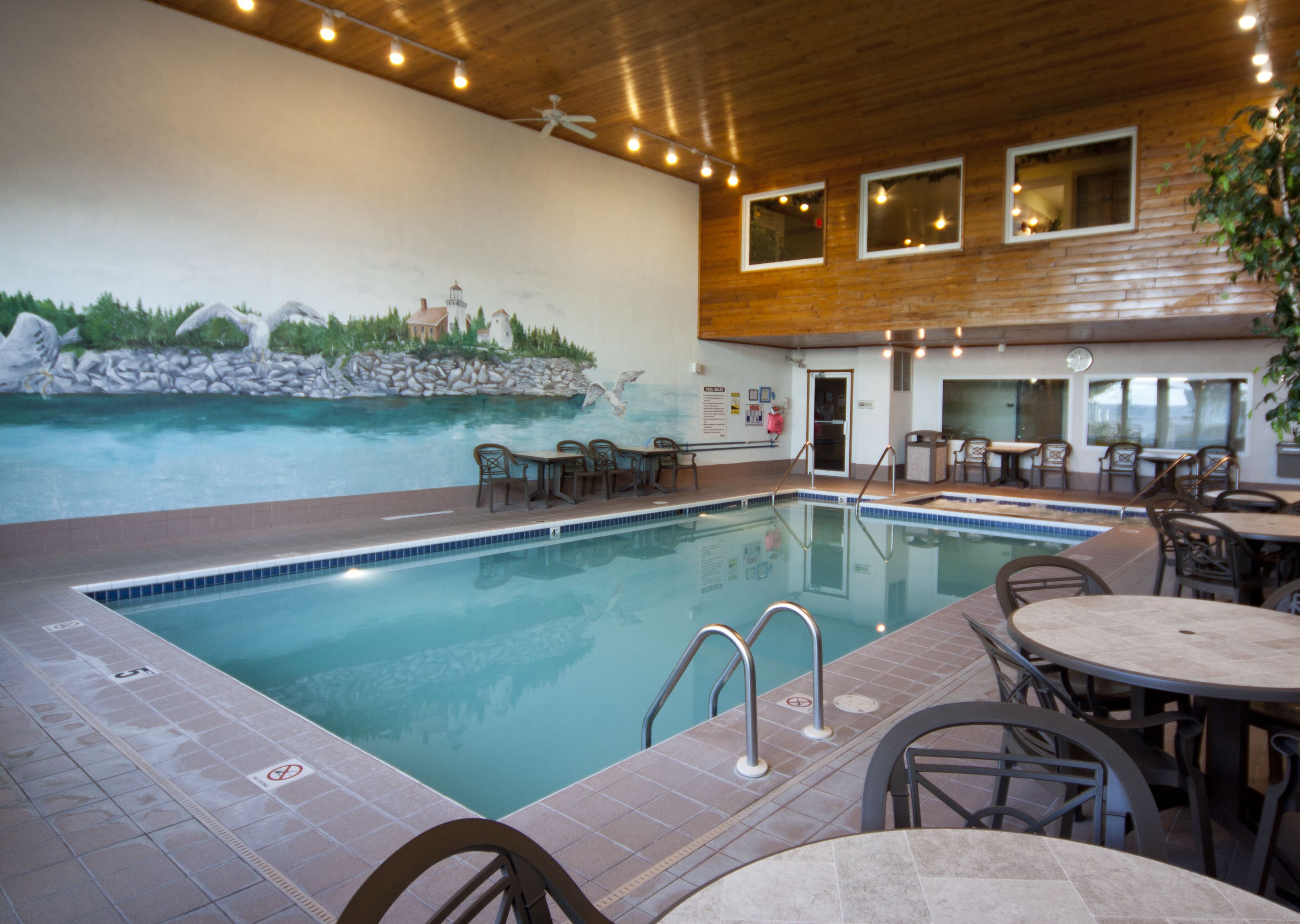 Una piscina interior con un mural de una escena costera, rodeada de mesas y sillas.