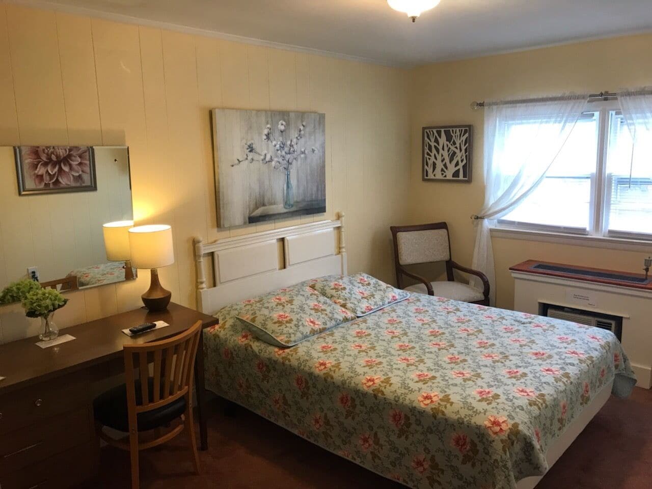 Une chambre à coucher avec un lit, un bureau, une chaise, une lampe et une fenêtre avec des rideaux.