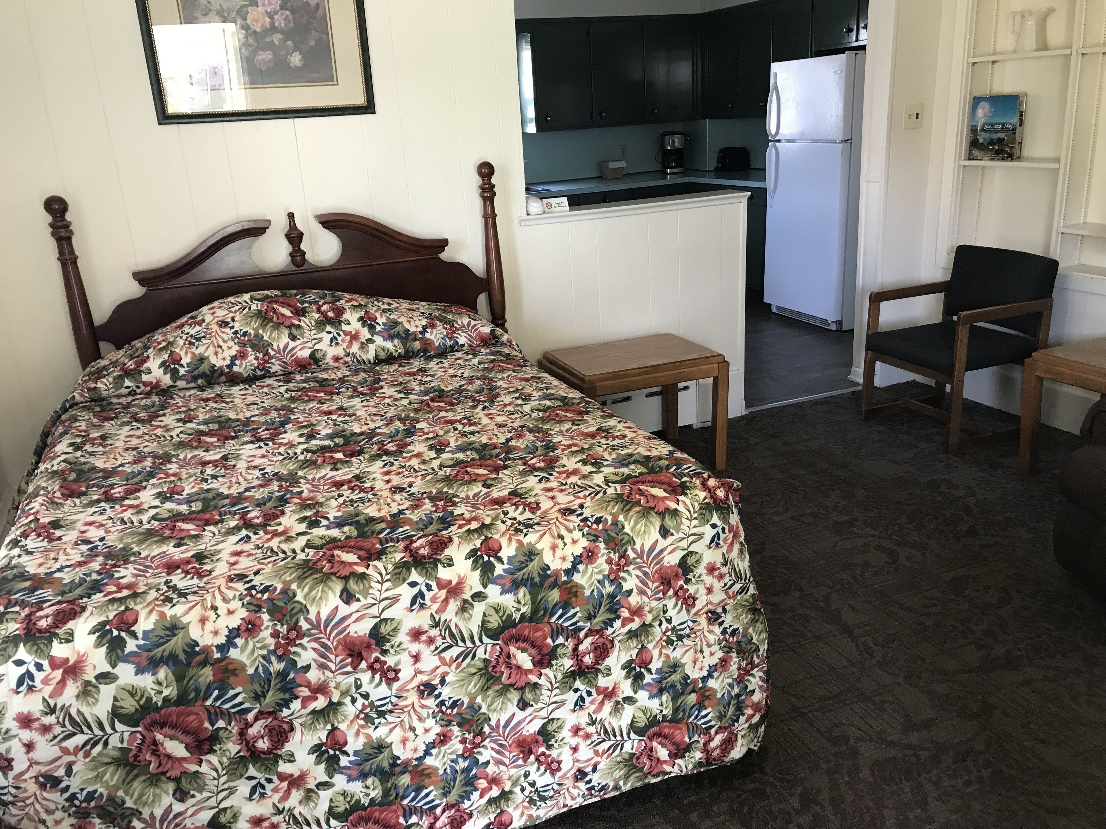 Un dormitorio con cubrecama floral, cabecera de madera, dos mesitas de noche y una silla. Se ve una kitchenette con refrigerador y armarios al fondo.
