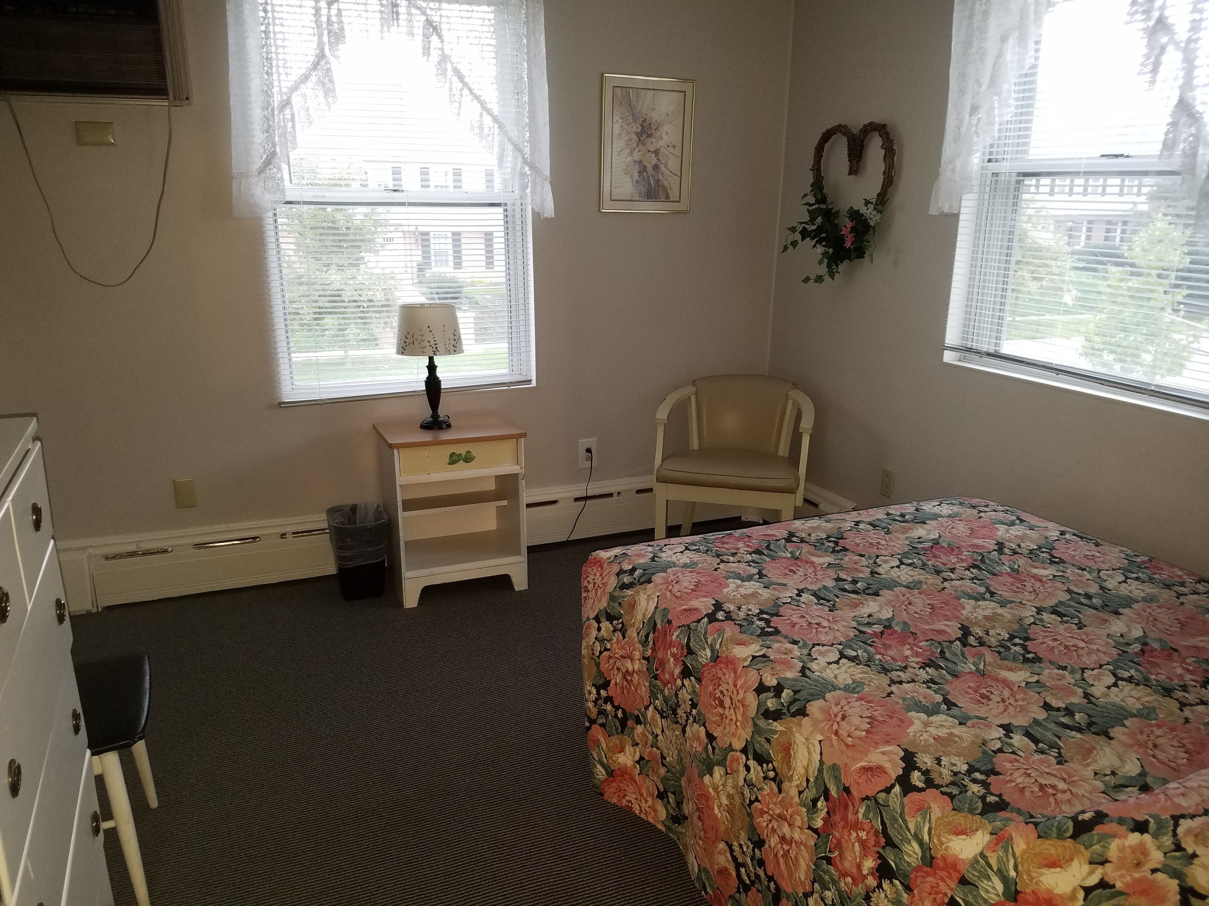 A bedroom with a floral bedspread, a chair, a nightstand with a lamp, and a picture on the wall.