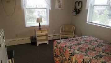 A bedroom with a floral bedspread, a chair, a nightstand with a lamp, and a picture on the wall.