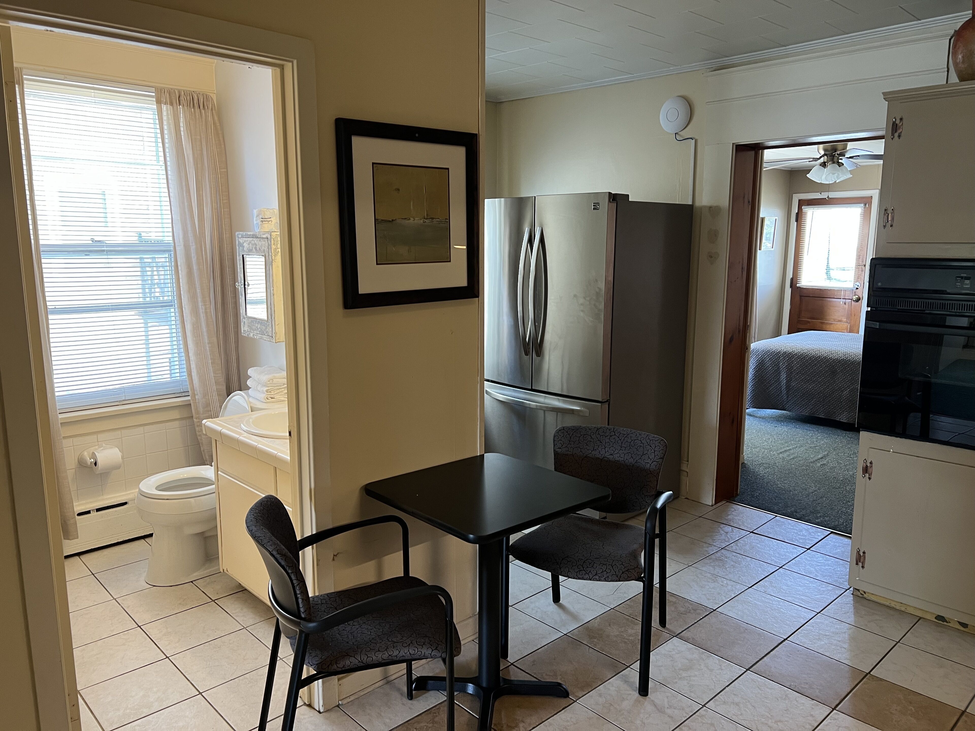 A compact kitchen with a refrigerator, a dining table with two chairs, a window with curtains, and a view of a bedroom through an open door.