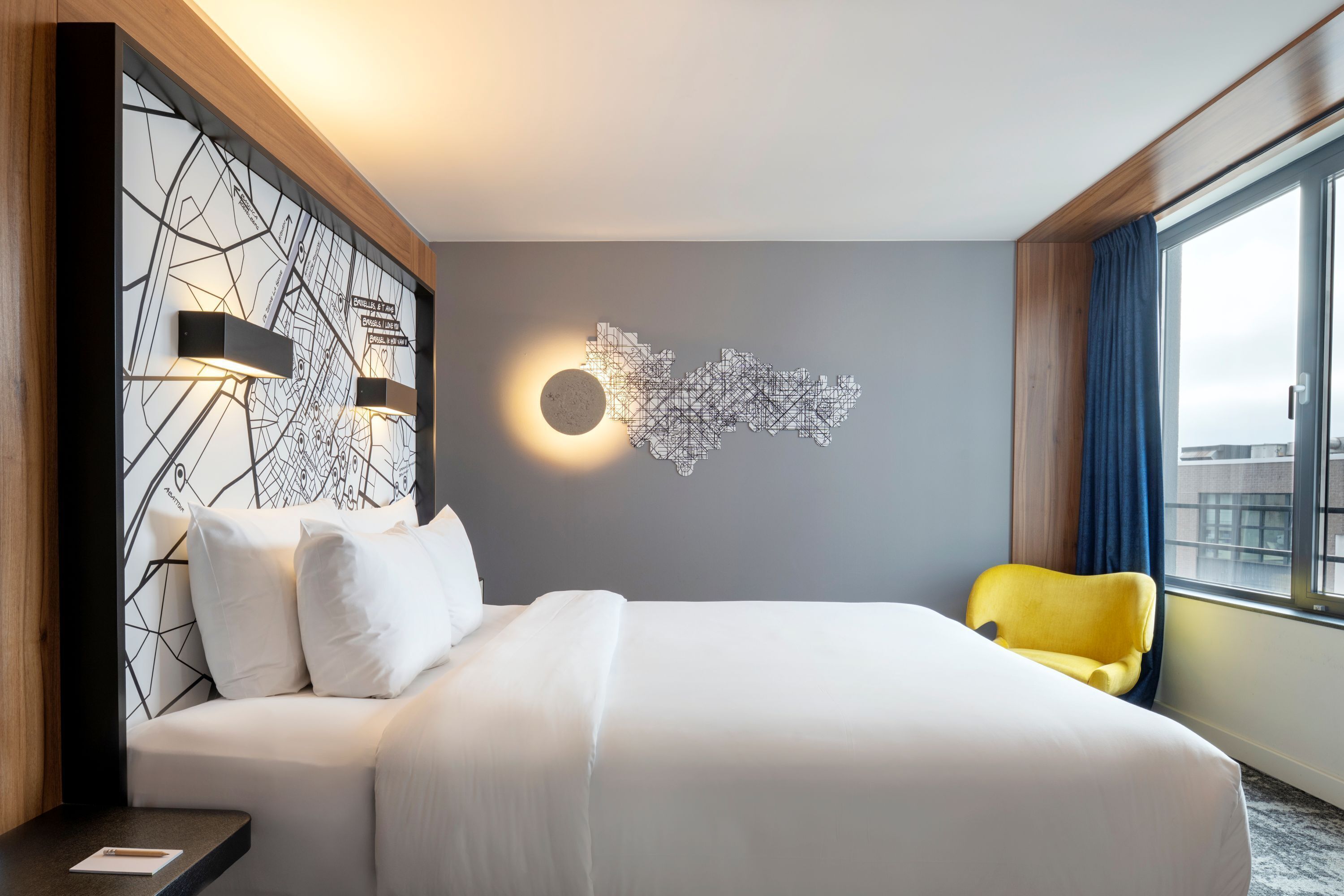 A hotel room with a large bed, a map wall art, a yellow chair, and a window with curtains.