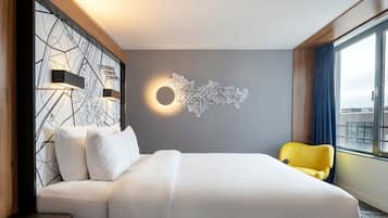 A hotel room with a large bed, a map wall art, a yellow chair, and a window with curtains.
