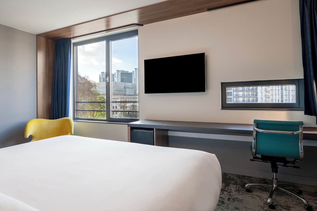 A hotel room with a bed, a desk, a chair, a TV, and a window with a city view.