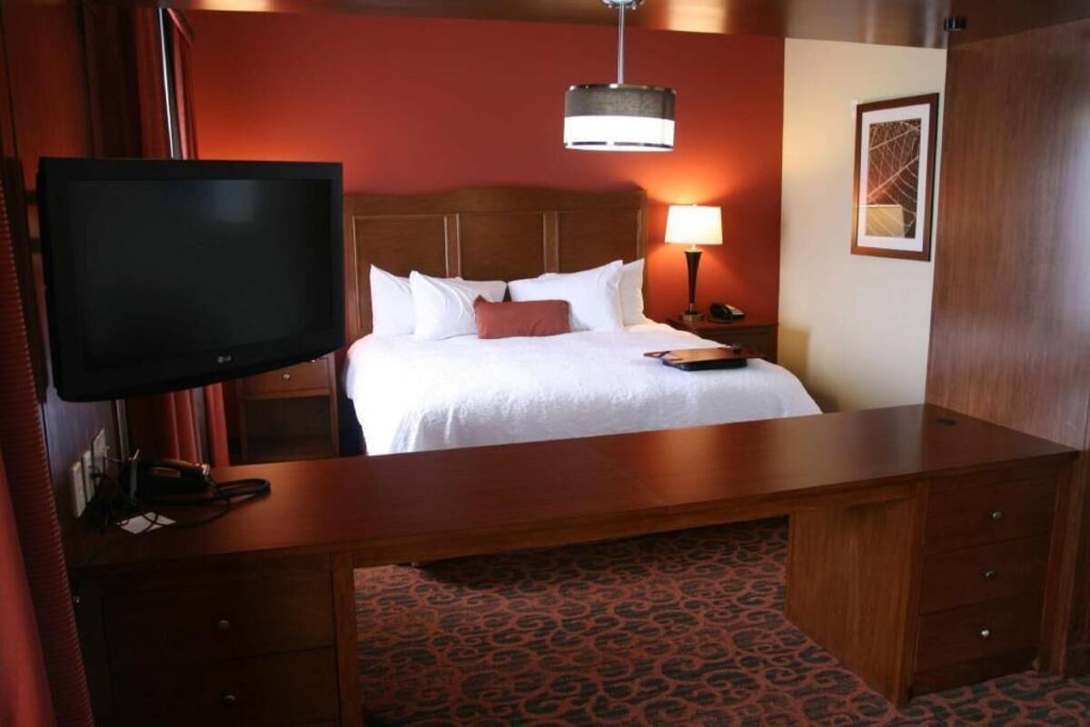 Habitación de hotel con cama, escritorio, televisor, lámpara y un cuadro en la pared.