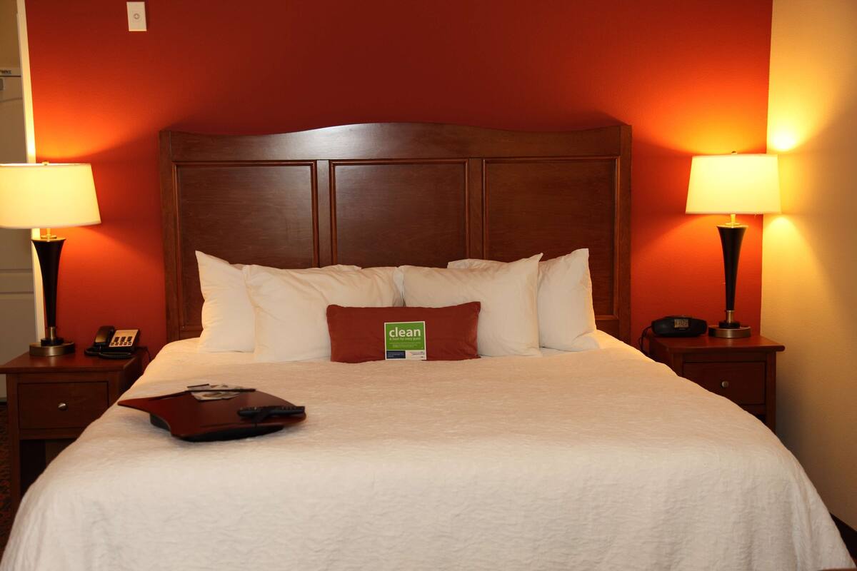 Habitación de hotel con una cama grande, dos mesitas de noche con lámparas, una pared roja y un cartel que indica que está limpia sobre la cama.