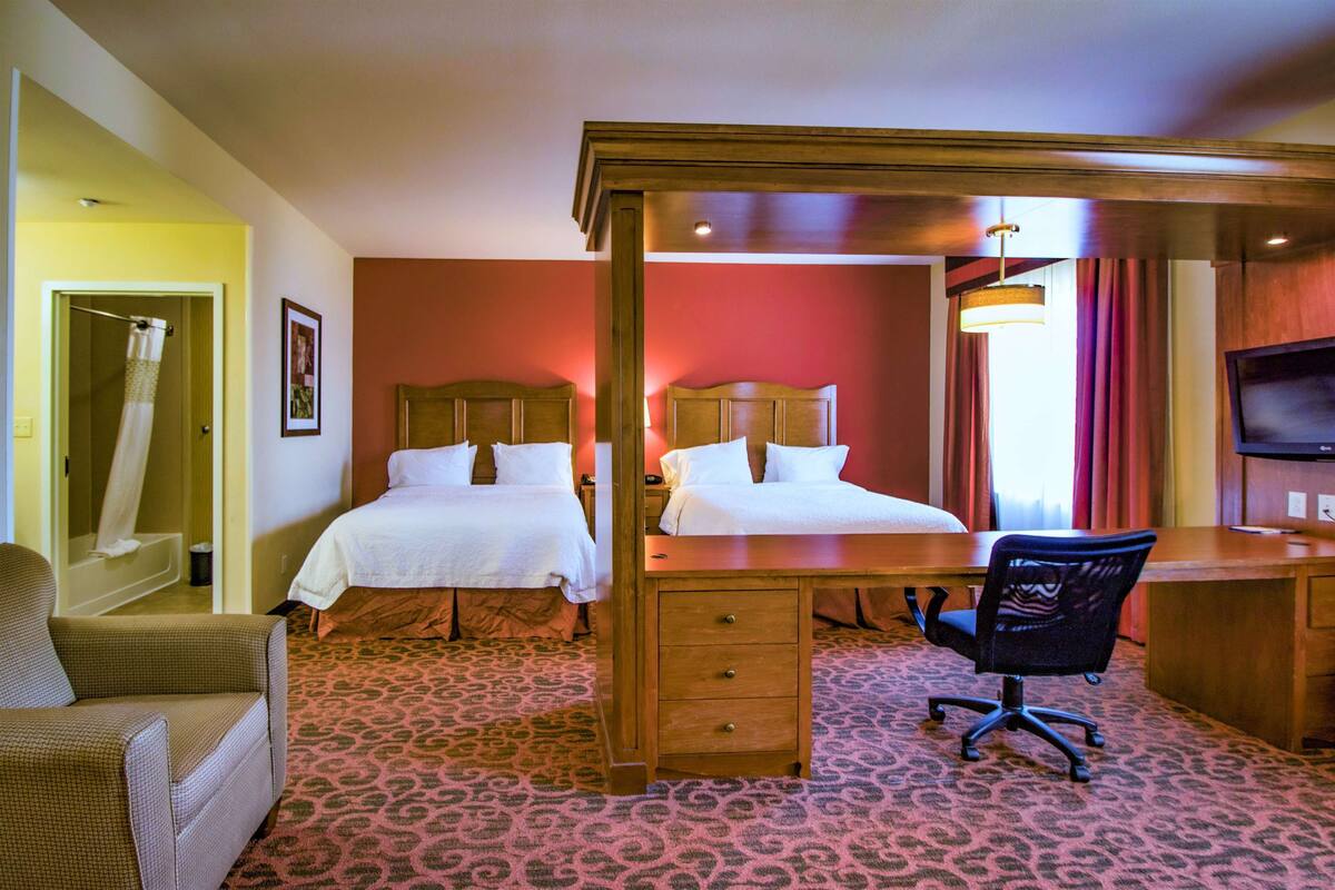 Habitación de hotel con dos camas, un escritorio, una silla, un televisor y un baño visible a través de una puerta abierta.