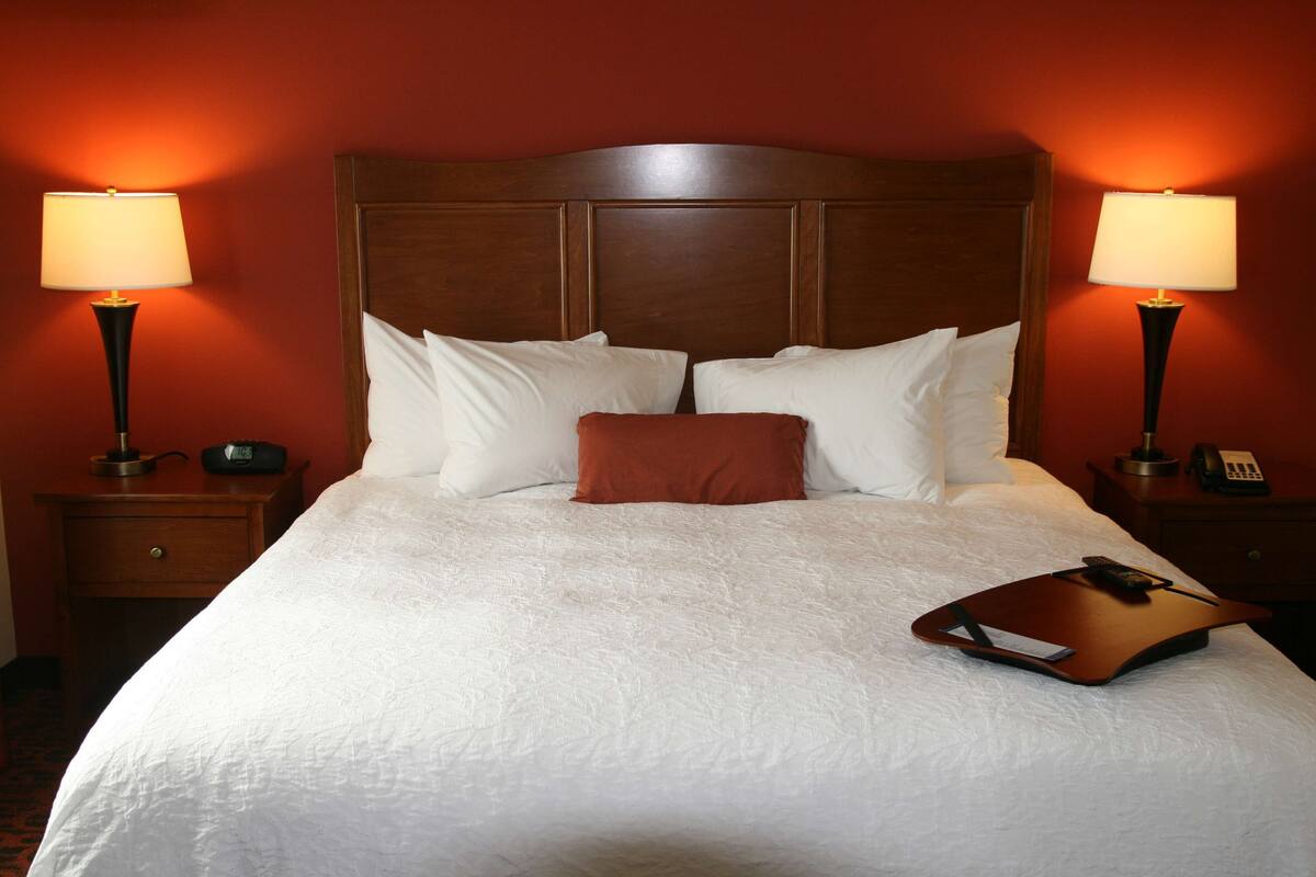 Una cama bien hecha con sábanas blancas y un almohadón marrón, flanqueada por dos mesitas de noche con lámparas, en una habitación con paredes rojas.
