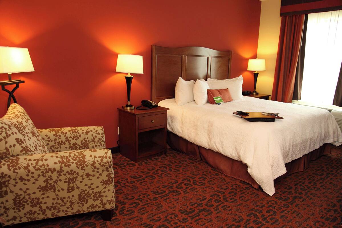 Una habitación de hotel con cama, dos lámparas de noche, teléfono, una silla y una alfombra estampada.