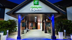 A Holiday Inn Express building entrance with a covered walkway, potted plants, and a bench. - Holiday Inn Express Colchester by IHG (Colchester)