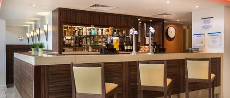Un espace bar avec un comptoir en bois, de nombreuses bouteilles d’alcool et une horloge au mur.