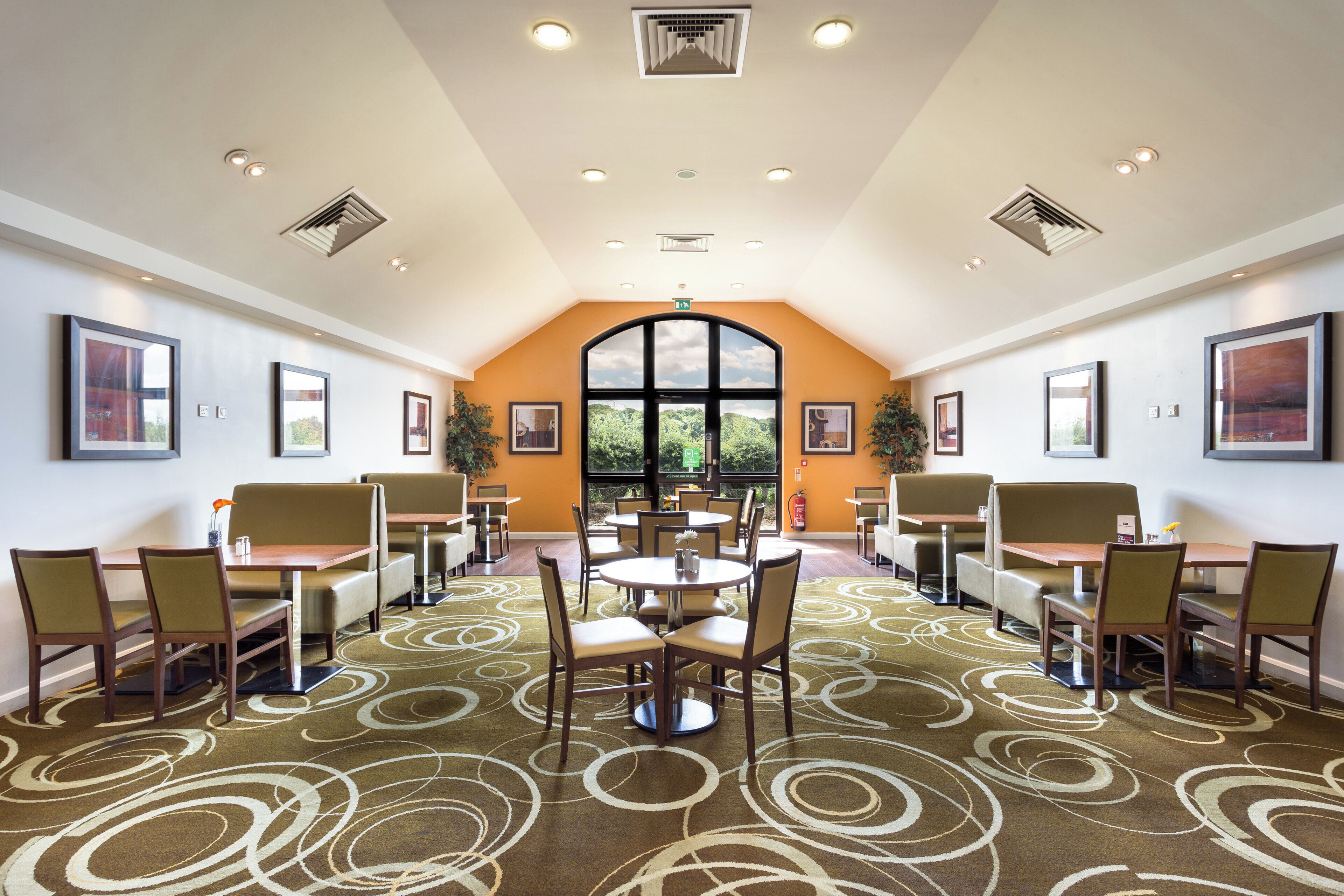 A spacious dining area with multiple tables and chairs, a large arched window with a view of greenery, and ceiling vents.